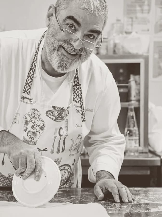 Gözlüklü, kır sakallı bir aşçı; beyaz ceketinin üstünde desenli önlük, mermer tezgahta bir tabağı tutarken gülümsüyor.