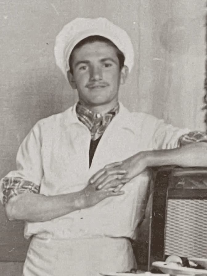 Eski bir baskıda genç bir aşçı: beyaz şapka ve ceket, boynunda desenli fular, koluyla bir sandalyenin arkasına yaslanmış.