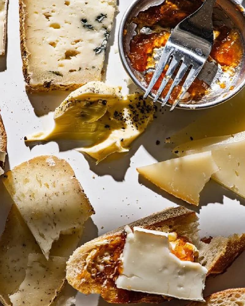 Beyaz zeminde kızarmış ekmek üstünde peynir, yanında çatalıyla metal kasede reçel ve bir dilim brie.
